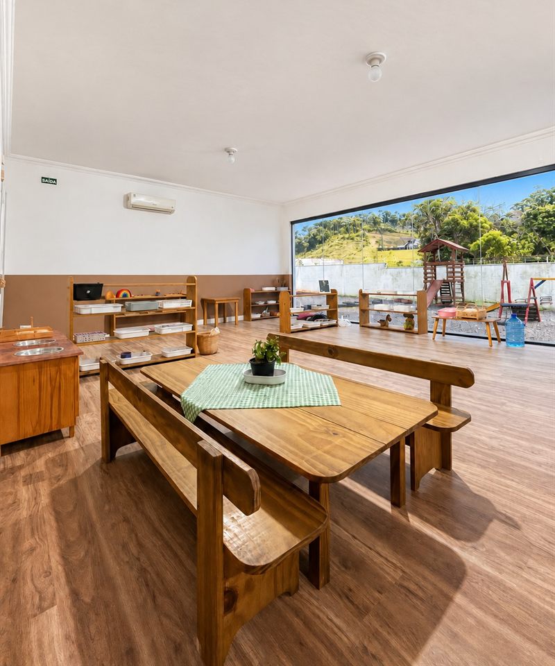 Sala do Programa Nido: ambiente amplo com mesa e bancos de madeira, prateleiras Montessori e janelão com vista para o parquinho